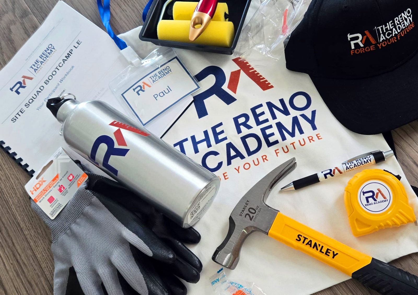 Site Squad Bootcamp LE enrolment kit flat-lay, Reno Academy workbook, water bottle, cap, name tag, work gloves, framing hammer, branded tape measure, utility knife and paint roller